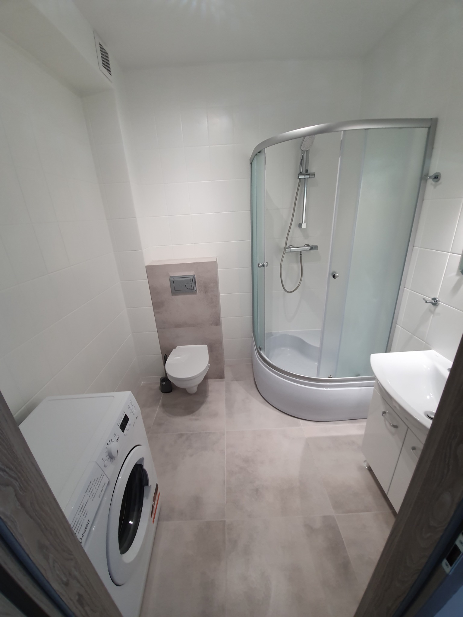 Jasna łazienka po remoncie z kabiną prysznicową, umywalką z szafką, toaletą podwieszaną i pralką Indesit.