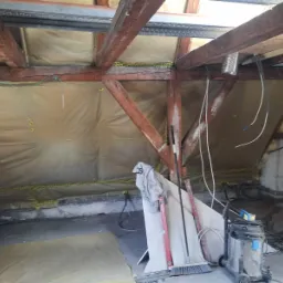 Poddasze w trakcie ocieplania wełną mineralną, widoczne drewniane krokwie, folia paroizolacyjna, poziomica, odkurzacz przemysłowy i przewody elektryczne.