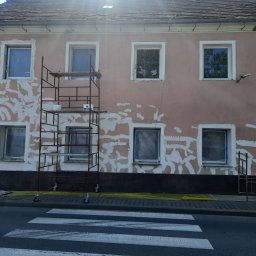 MaDrok - Elewacja budynku w trakcie przygotowania do prac wykończeniowych, z widocznymi śladami po szpachlowaniu i zabezpieczonymi oknami, rusztowanie przy ścianie, widok z poziomu ulicy.