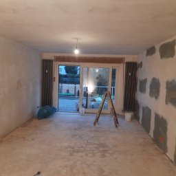 MaDrok - Pokój w trakcie remontu: ściany częściowo wyszpachlowane, widoczna drabina, grzejniki i worki z materiałami budowlanymi.