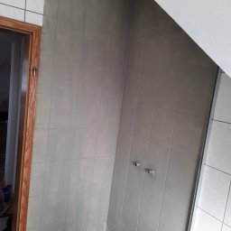 MaDrok - Przygotowanie ścian w łazience do remontu: szare płytki częściowo pokryte farbą, widoczne przyłącza wodne i zabezpieczona podłoga folią malarską.