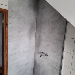 MaDrok - Szara ściana w kabinie prysznicowej z zamontowaną baterią, imitująca beton, przylegająca do białych płytek i skośnego sufitu.