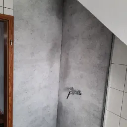 Szara ściana w kabinie prysznicowej z zamontowaną baterią, imitująca beton, przylegająca do białych płytek i skośnego sufitu.