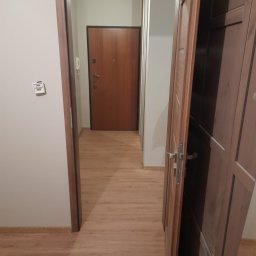 MaDrok - Widok na korytarz z panelami podłogowymi w odcieniach brązu, z otwartymi drewnianymi drzwiami po prawej stronie i drzwiami wejściowymi w tle.