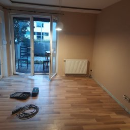 MaDrok - Pokój po remoncie z ułożonymi panelami podłogowymi, widoczne narzędzia pracy i otwarte drzwi balkonowe.