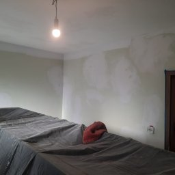 MaDrok - Pokój w trakcie przygotowań do malowania: ściany z nałożonymi punktowo łatami szpachli, podłoga zabezpieczona czarną folią, na folii czerwony ręcznik, widoczna żarówka na kablu.