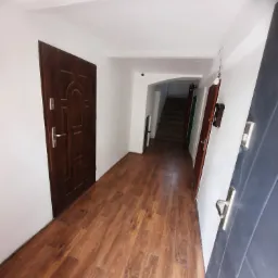 Korytarz po remoncie z nową podłogą z paneli imitujących drewno, świeżo pomalowane ściany na biało, widoczne drzwi wejściowe w kolorze ciemnego brązu i schody w tle.