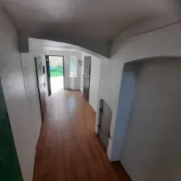 Korytarz z brązowymi panelami podłogowymi, świeżo otynkowanymi ścianami i dwiema parami drzwi po bokach, z widokiem na zewnątrz przez drzwi wejściowe.