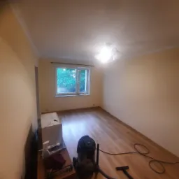 Pomalowane na beżowo wnętrze pokoju z drewnianą podłogą po remoncie, widoczny odkurzacz przemysłowy i narzędzia malarskie, okno z widokiem na zieleń.