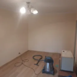 Pusta, świeżo pomalowana beżowa ściana w pokoju z ułożonymi panelami podłogowymi, widoczny odkurzacz przemysłowy i osłonięty mebel lub urządzenie, sufit z lampą.