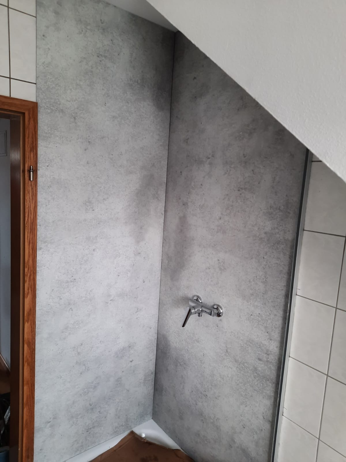 Szara ściana w kabinie prysznicowej z zamontowaną baterią, imitująca beton, przylegająca do białych płytek i skośnego sufitu.