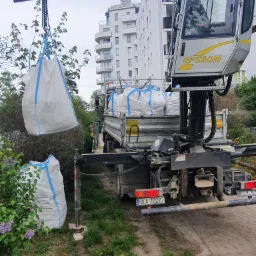 Żuraw przenosi duży biały worek z zawartością na teren budowy z zielenią w tle, w tle widoczny budynek mieszkalny.