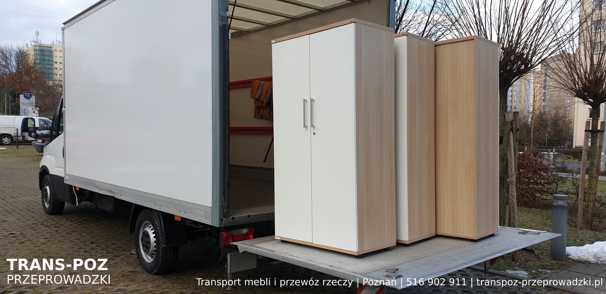 Trzy szafy na platformie załadunkowej białej ciężarówki przeprowadzkowej na tle miejskiego krajobrazu z budynkami.