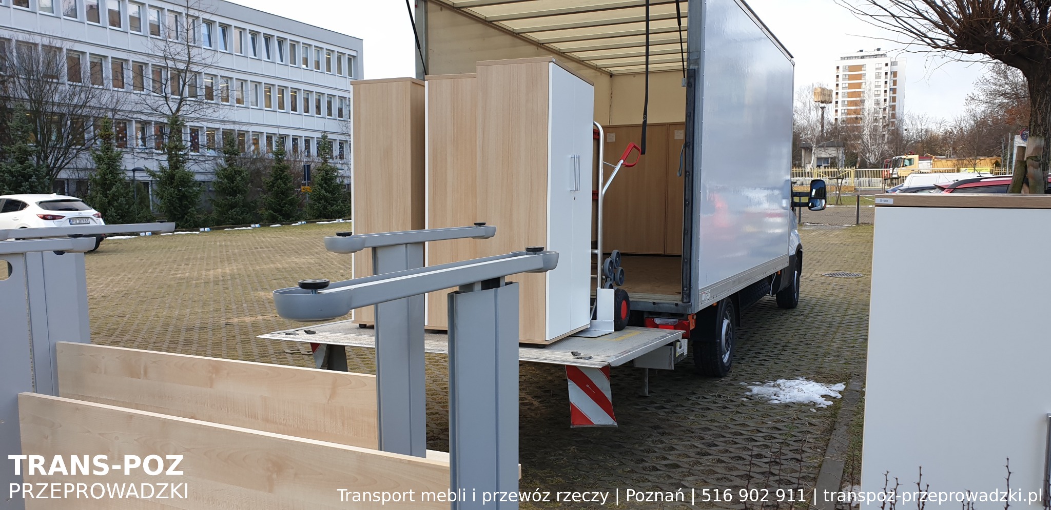 Meble częściowo załadowane na platformę windy załadowczej białej ciężarówki, budynki w tle, widoczne logo firmy przeprowadzkowej.