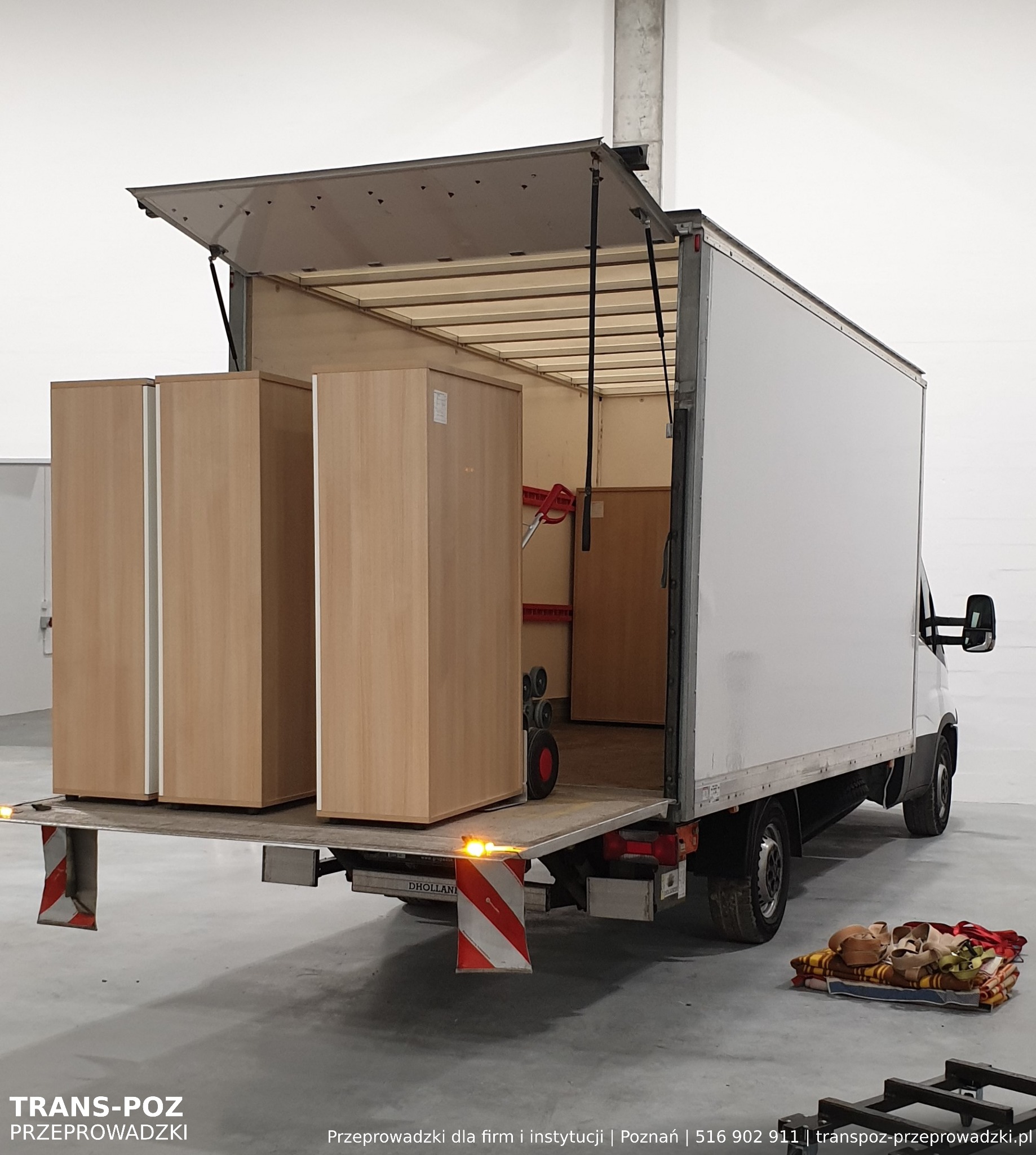 Dwa drewniane regały załadowane na platformę windy samochodu dostawczego, przygotowane do transportu, z widocznymi pasami i kocami zabezpieczającymi na podłodze.