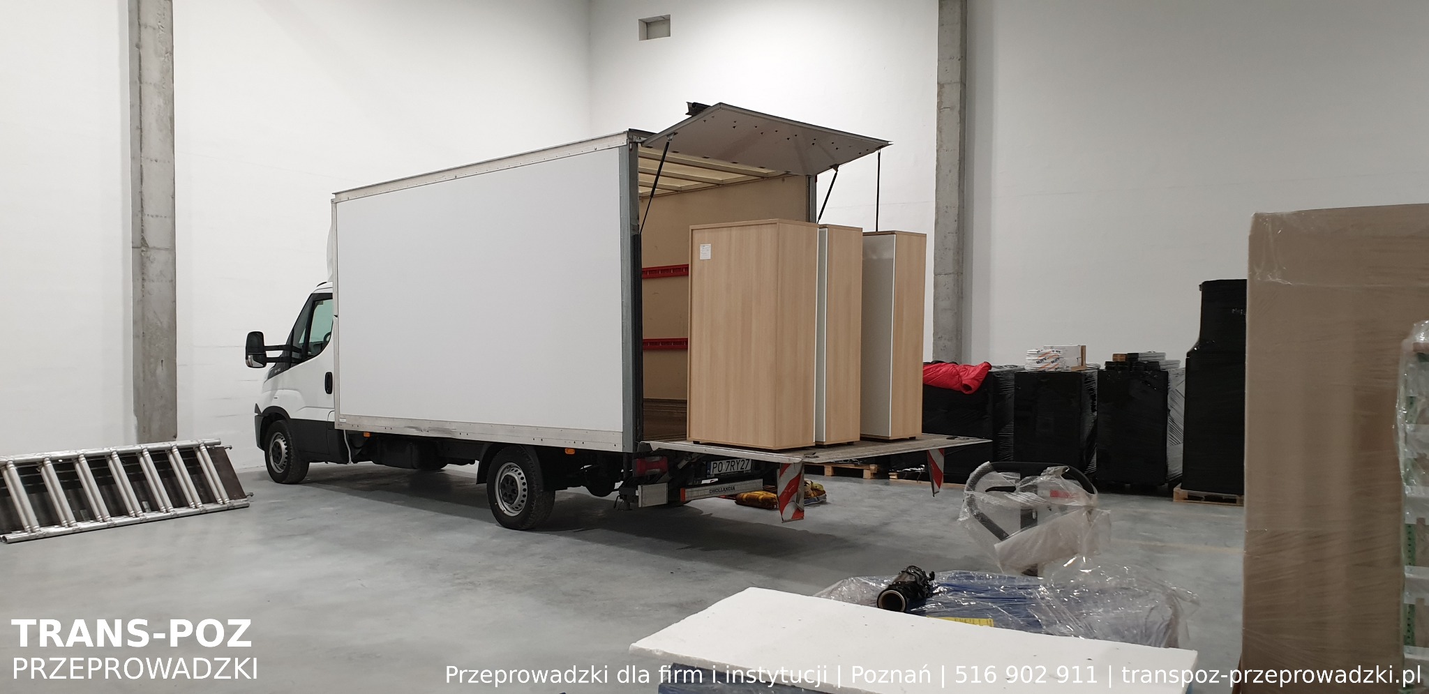 Dwa jasne, drewniane biurka w otwartym bagażniku białego busa dostawczego podczas załadunku w magazynie, drabina oparta o bok pojazdu.