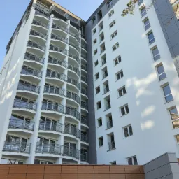 Wysoki budynek mieszkalny z licznymi balkonami z metalowymi balustradami i elewacją w odcieniach bieli i szarości, widziany z dołu pod ostrym kątem.