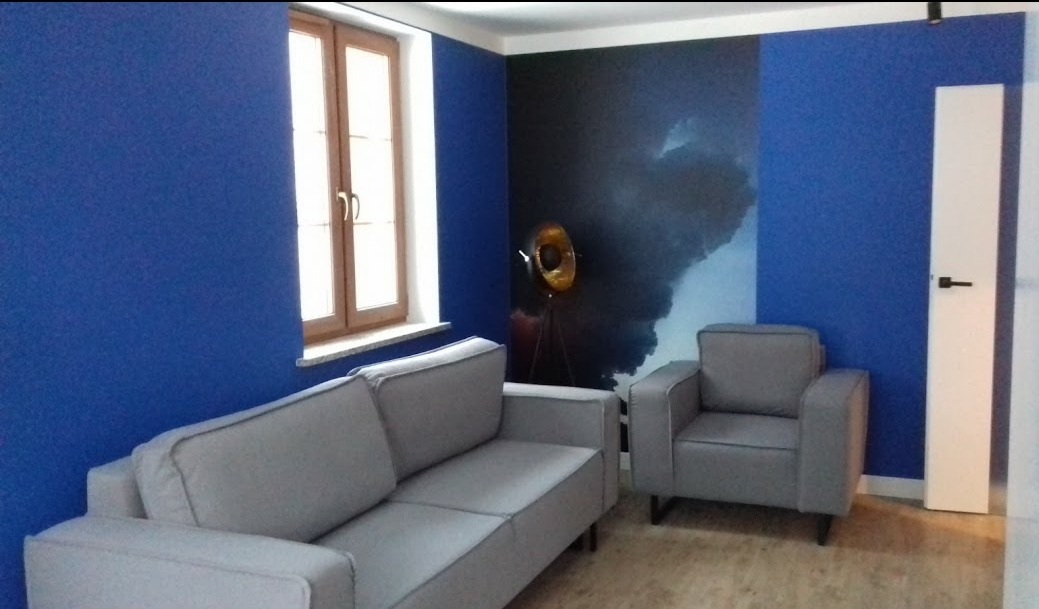 Niebieskie ściany, szara sofa i fotel w pokoju z drewnianym oknem i lampą na tle tapety imitującej burzę.