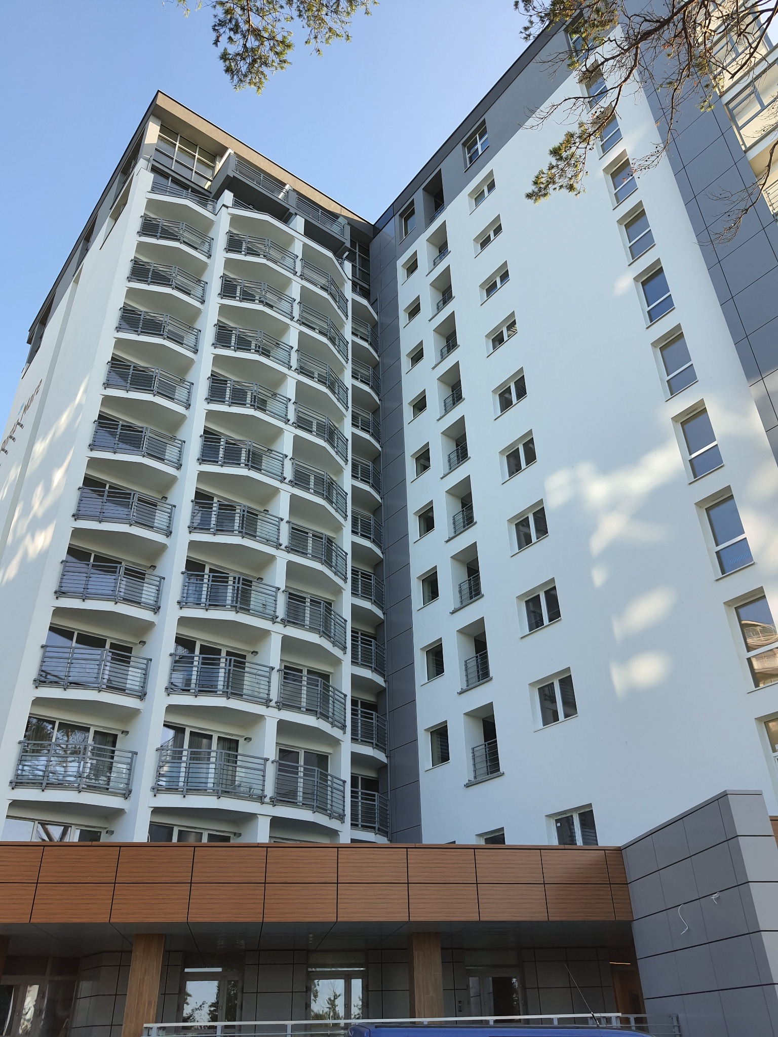 Wysoki budynek mieszkalny z licznymi balkonami z metalowymi balustradami i elewacją w odcieniach bieli i szarości, widziany z dołu pod ostrym kątem.