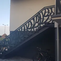 Zewnętrzne schody metalowe z ażurową balustradą o motywie roślinnym, prowadzące do wejścia na piętro budynku, z rowerem zaparkowanym pod spodem.