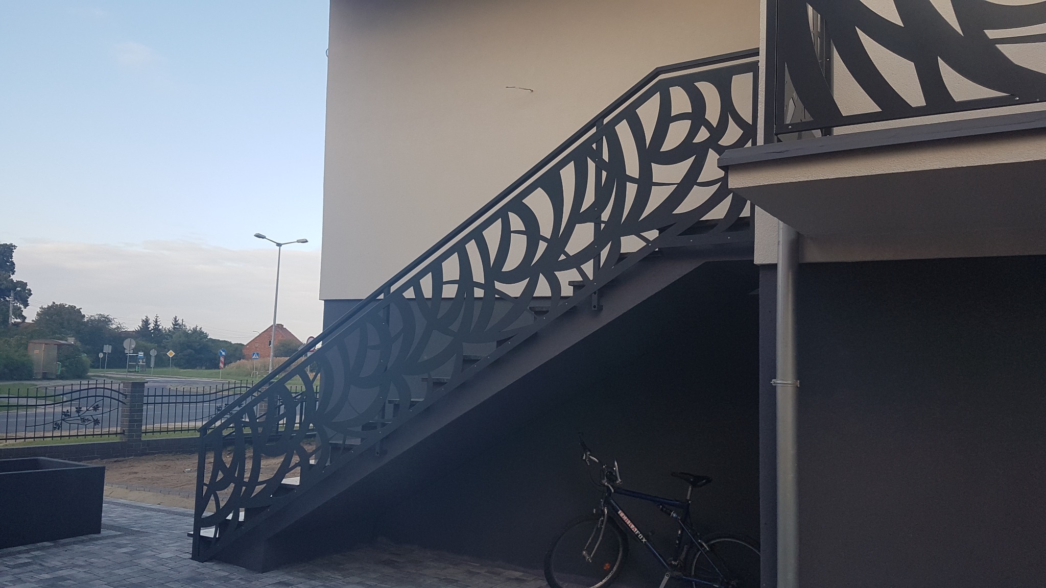 Zewnętrzne schody metalowe z ażurową balustradą o motywie roślinnym, prowadzące do wejścia na piętro budynku, z rowerem zaparkowanym pod spodem.