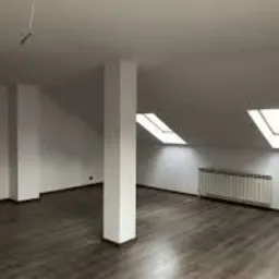 Puste, jasne poddasze z drewnianą podłogą, białymi ścianami, kolumną konstrukcyjną i dwoma oknami dachowymi; grzejnik pod oknem.