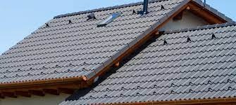 Szary dach z dachówki ceramicznej z widocznym oknem dachowym i systemem rynnowym z drewna, na tle błękitnego nieba.