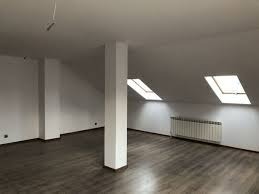 Puste, jasne poddasze z drewnianą podłogą, białymi ścianami, kolumną konstrukcyjną i dwoma oknami dachowymi; grzejnik pod oknem.