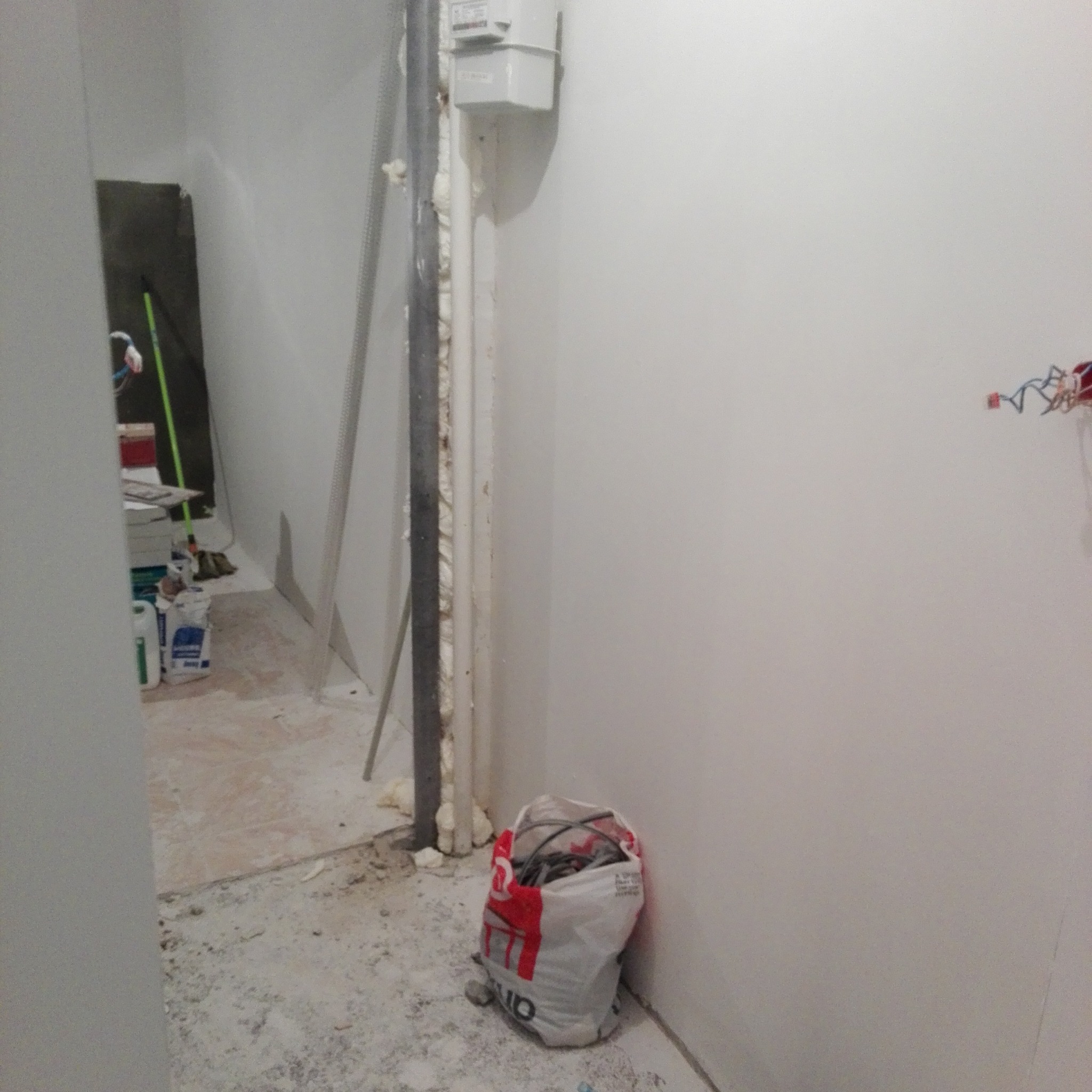 Wnętrze w trakcie prac wykończeniowych: widoczne rury osadzone pianką montażową, listwa przypodłogowa, worki z materiałami budowlanymi oraz porozrzucane resztki gruzu i pyłu na podłodze.