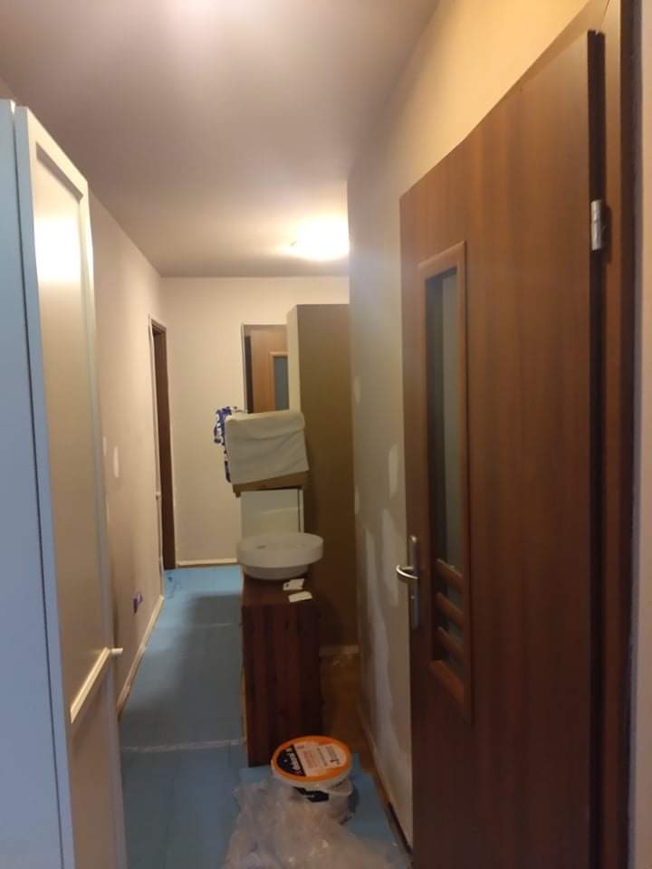 Korytarz w trakcie odświeżania: podłoga zabezpieczona folią, ściany przygotowane do malowania, widoczny pojemnik z farbą i umywalka tymczasowo zabezpieczona folią malarską.