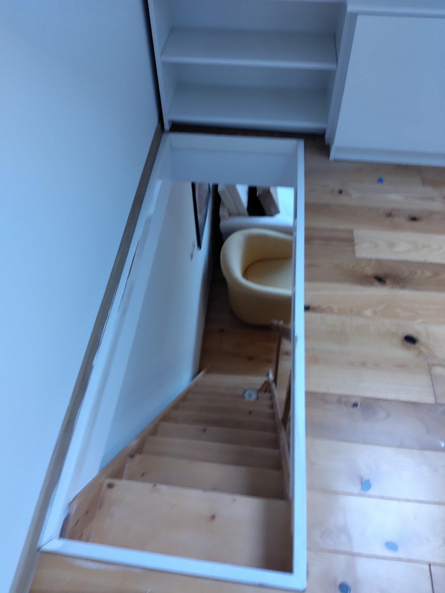 Ukryte schody do piwnicy pod zabudową meblową, widoczne drewniane stopnie i fragment jasnego fotela w dolnej części.