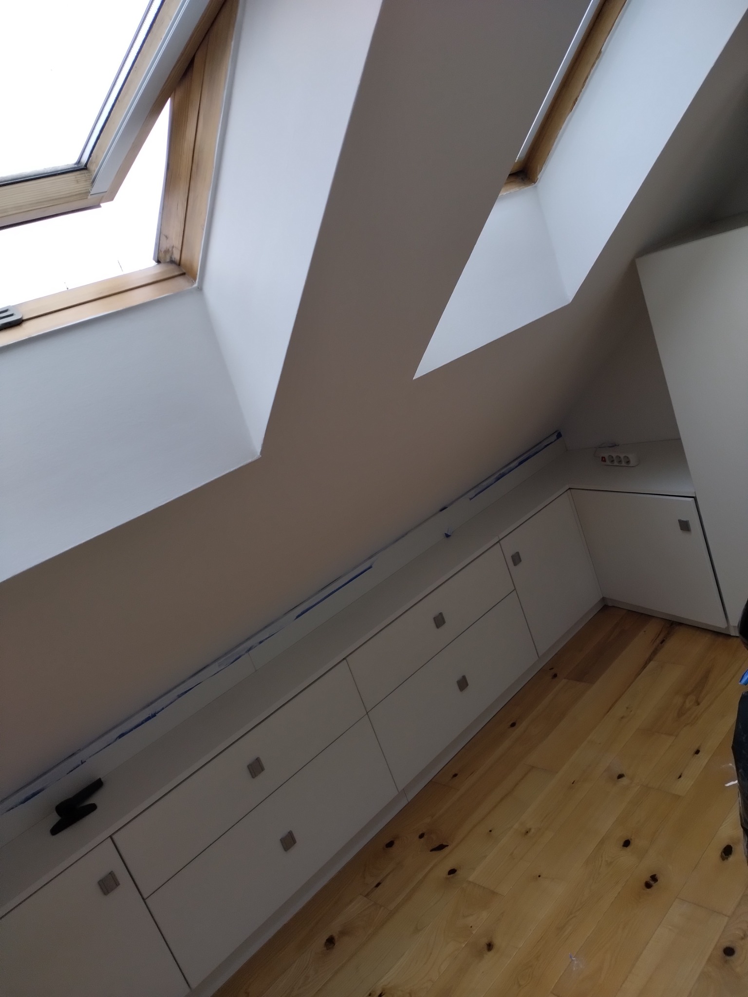Białe szafki w zabudowie pod skosem na poddaszu z drewnianą podłogą, widoczne okna dachowe i listwa maskująca.