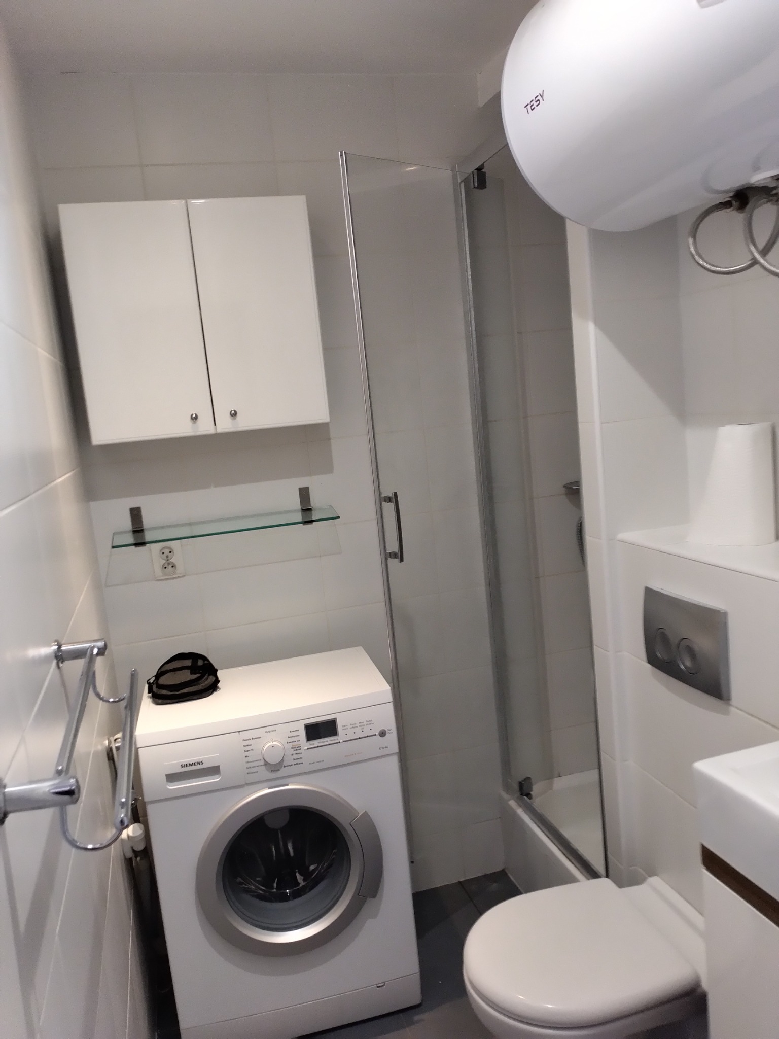 Mała, biała łazienka z kabiną prysznicową, pralką Siemens, wiszącą szafką, bojlerem Tesy i toaletą.
