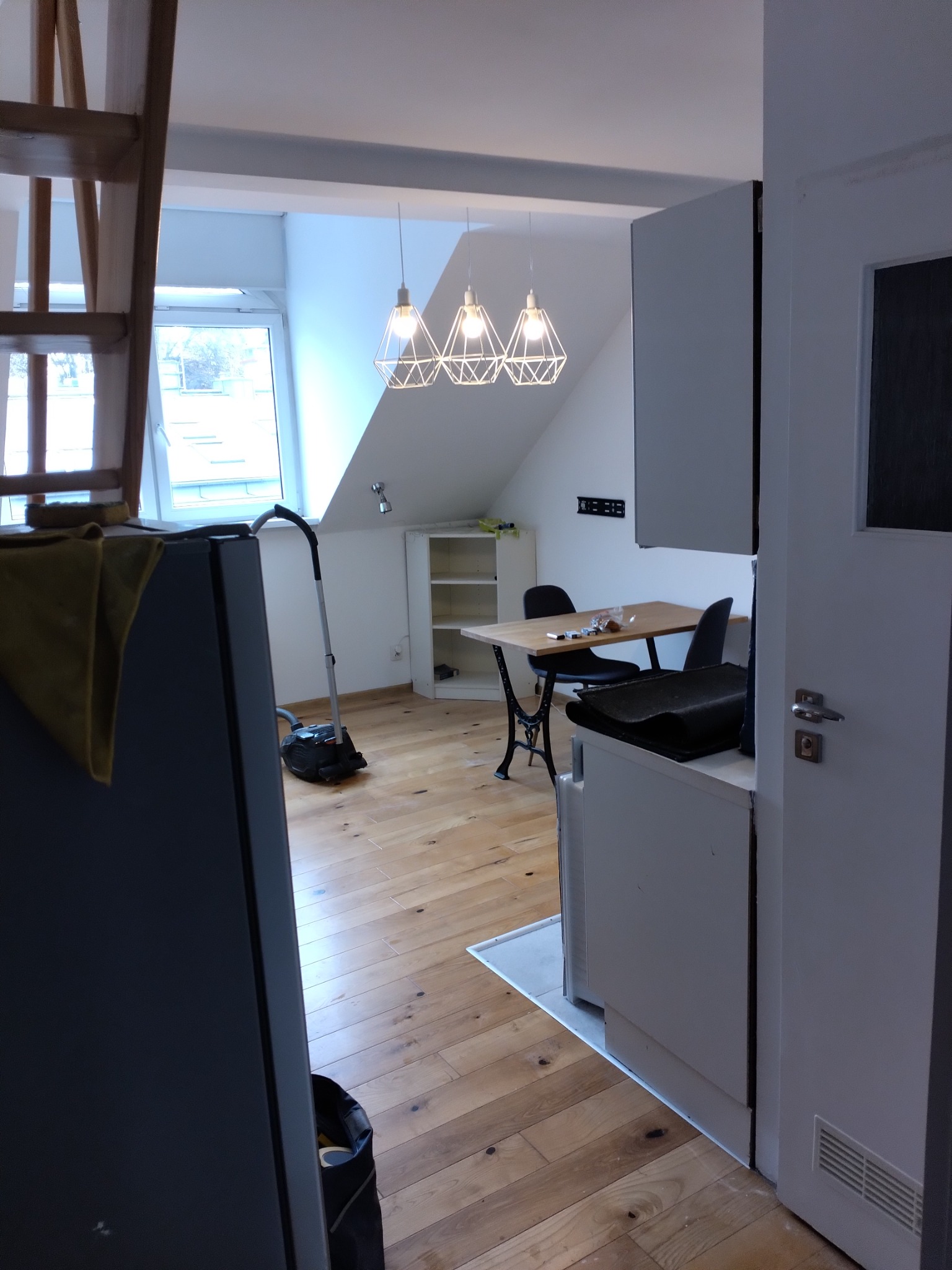 Wnętrze mieszkania po remoncie: drewniana podłoga, białe ściany, nowoczesne lampy wiszące, odkurzacz w rogu, stół z krzesłami i fragment kuchni z białymi szafkami.