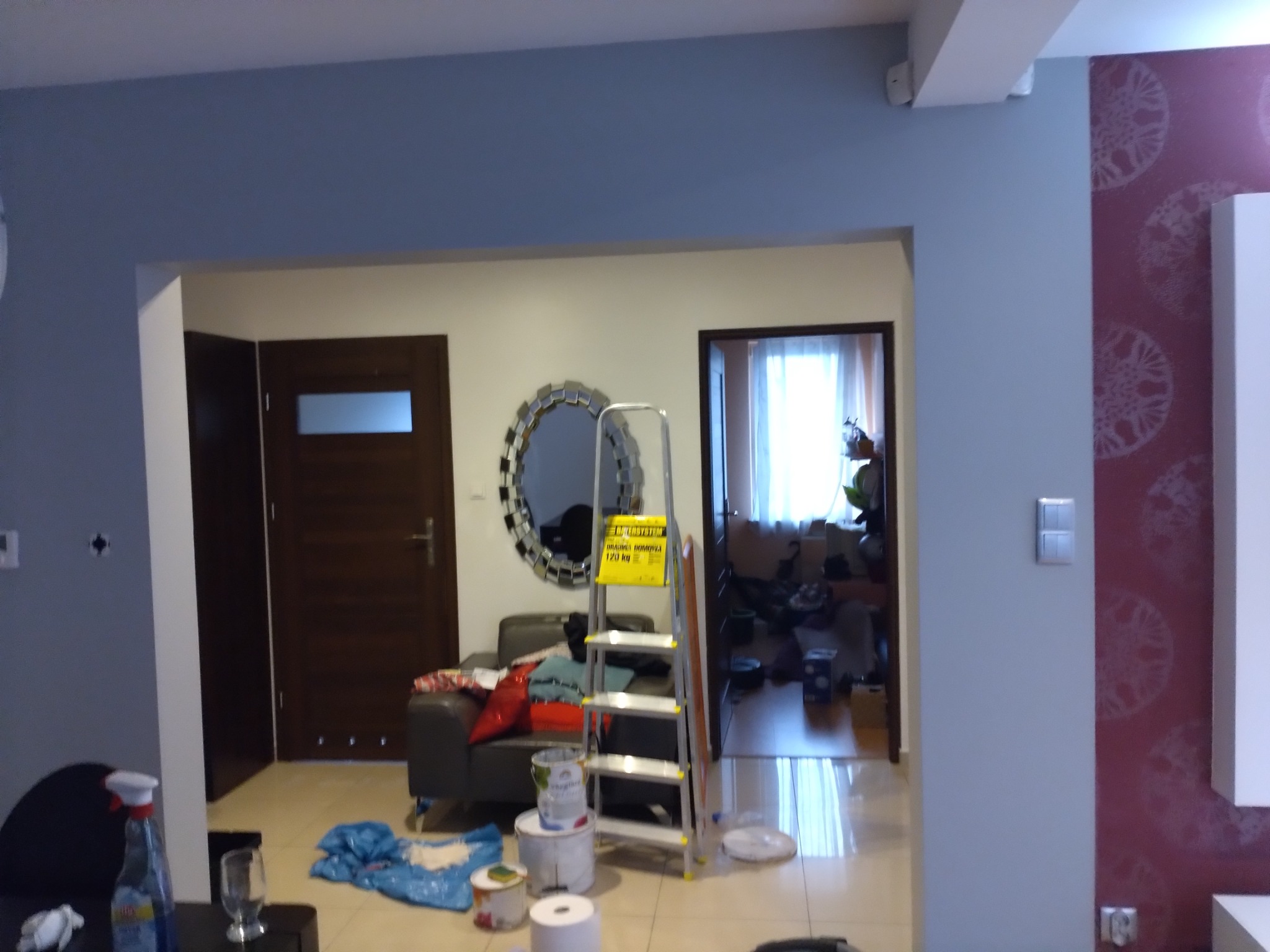 Wnętrze w trakcie odświeżania: drabina, puszki z farbą, folia malarska, ściany częściowo pomalowane na szaro-niebieski kolor, widoczne meble i dekoracje.