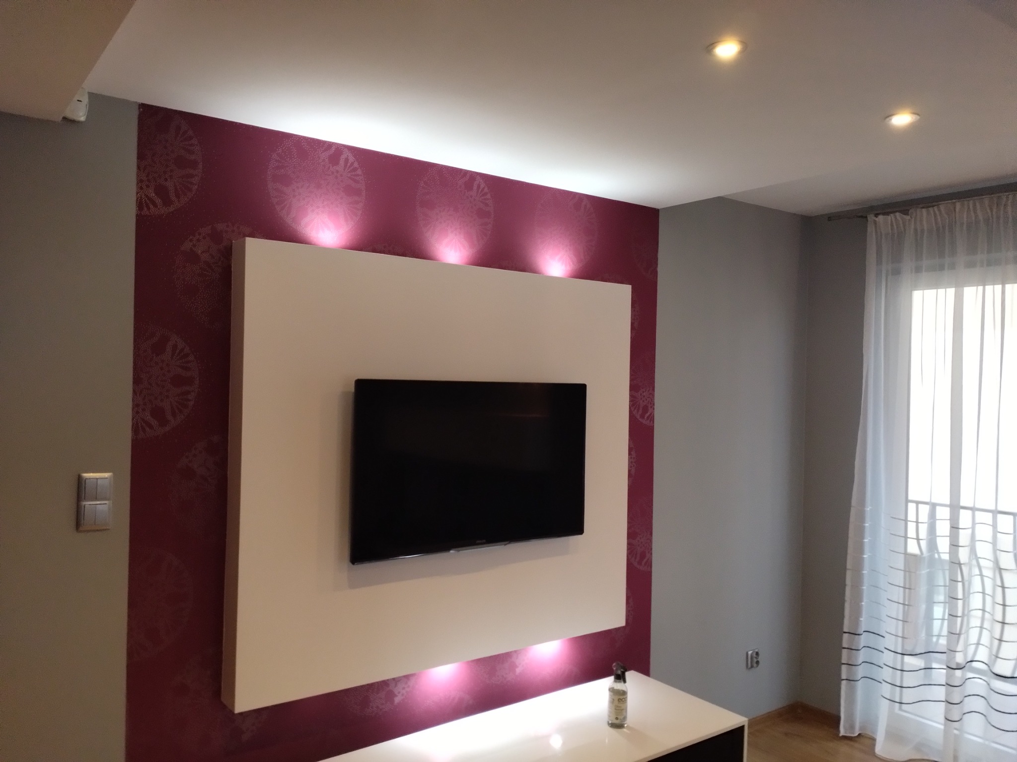 Nowoczesny salon z bordową ścianą w ornamenty, podświetlony panel pod telewizor, białe zasłony i parkiet na podłodze.