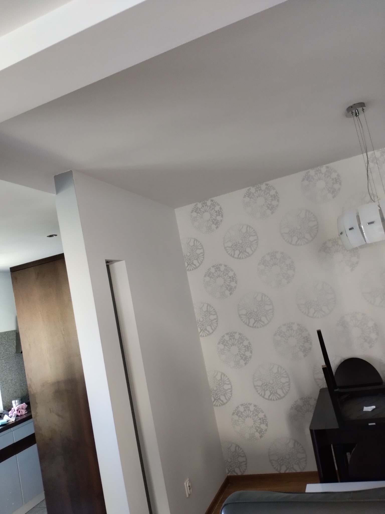 Białe ściany z wzorzystą tapetą w okrągłe ornamenty, nowoczesna lampa wisząca, widok na sąsiednie pomieszczenie z zabudową kuchenną.