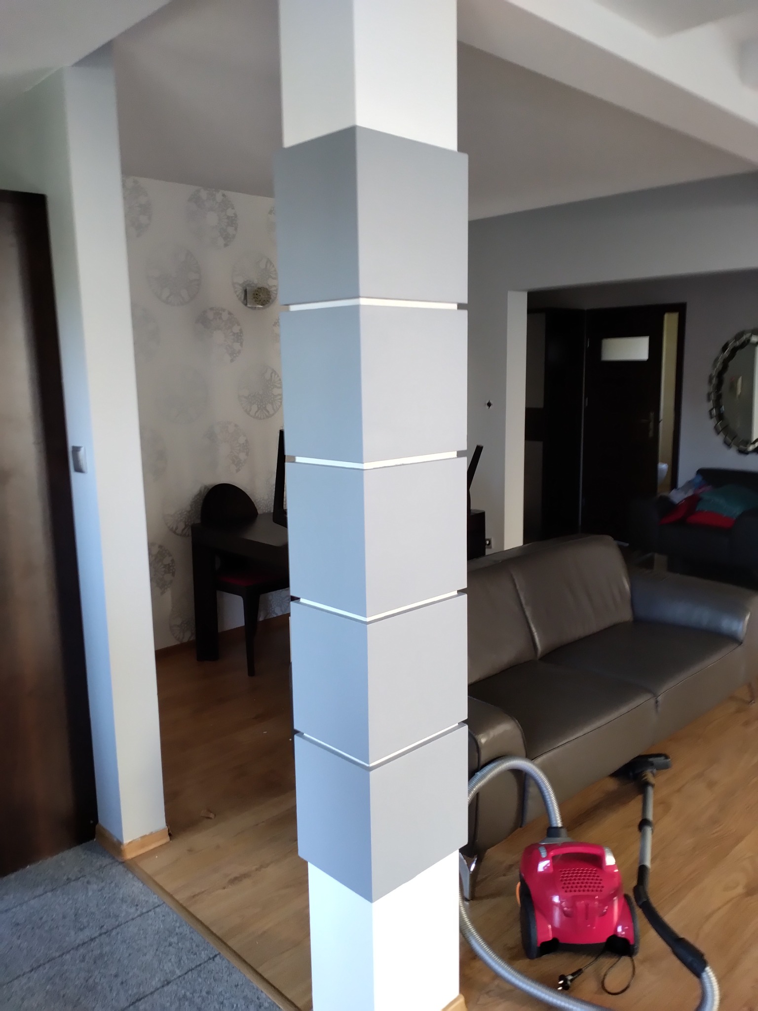 Szary filar dekoracyjny z białymi akcentami w nowoczesnym salonie, z widoczną częścią sofy, stołu i krzesła w tle, oraz odkurzaczem na pierwszym planie.