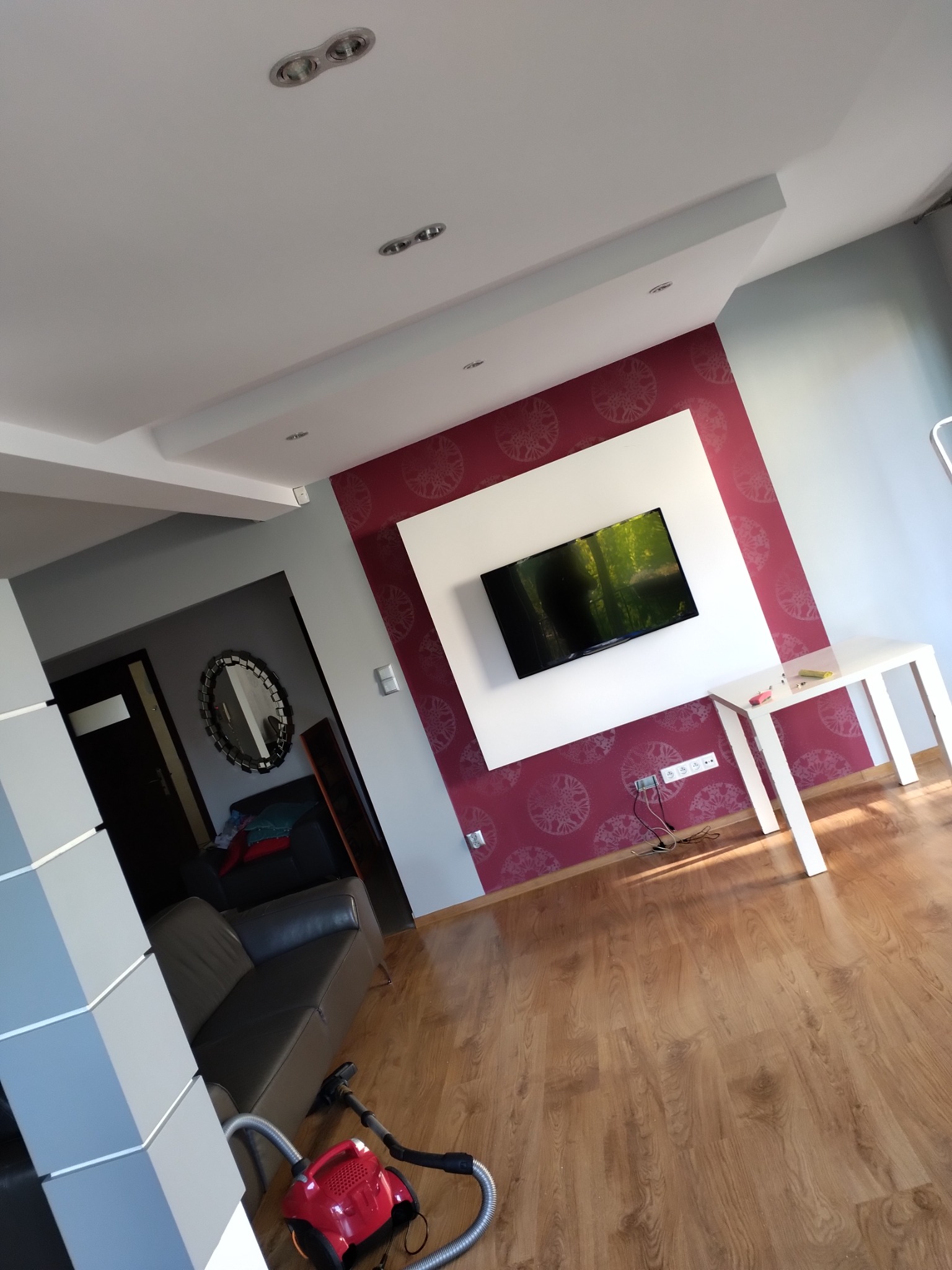 Salon po odświeżeniu z bordową ścianą z wzorem, białą ramą wokół telewizora, drewnianą podłogą i odkurzaczem w rogu.