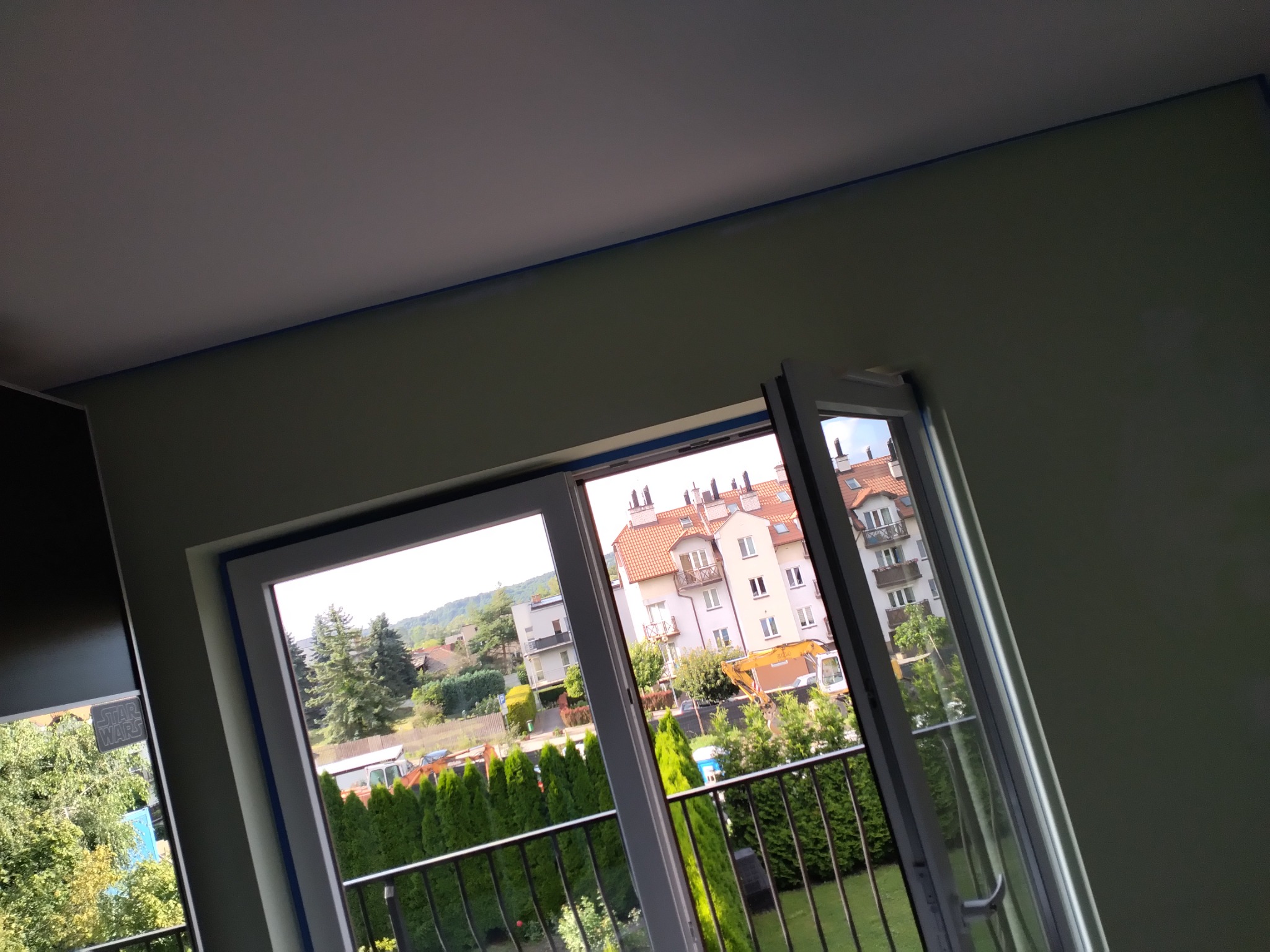 Świeżo pomalowany pokój z otwartym balkonem, widok na zieleń i budynki z czerwonymi dachami, zabezpieczone niebieską taśmą malarską.