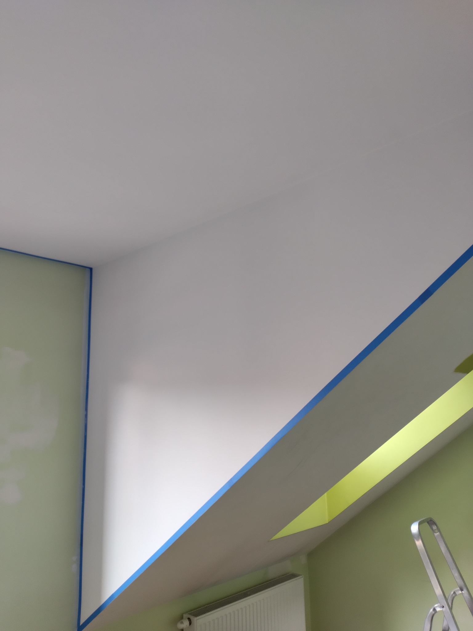 Przygotowanie do malowania: ściana i sufit zabezpieczone niebieską taśmą malarską, widoczny fragment drabiny i grzejnika, ściana pomalowana na jasnozielono, sufit na biało.