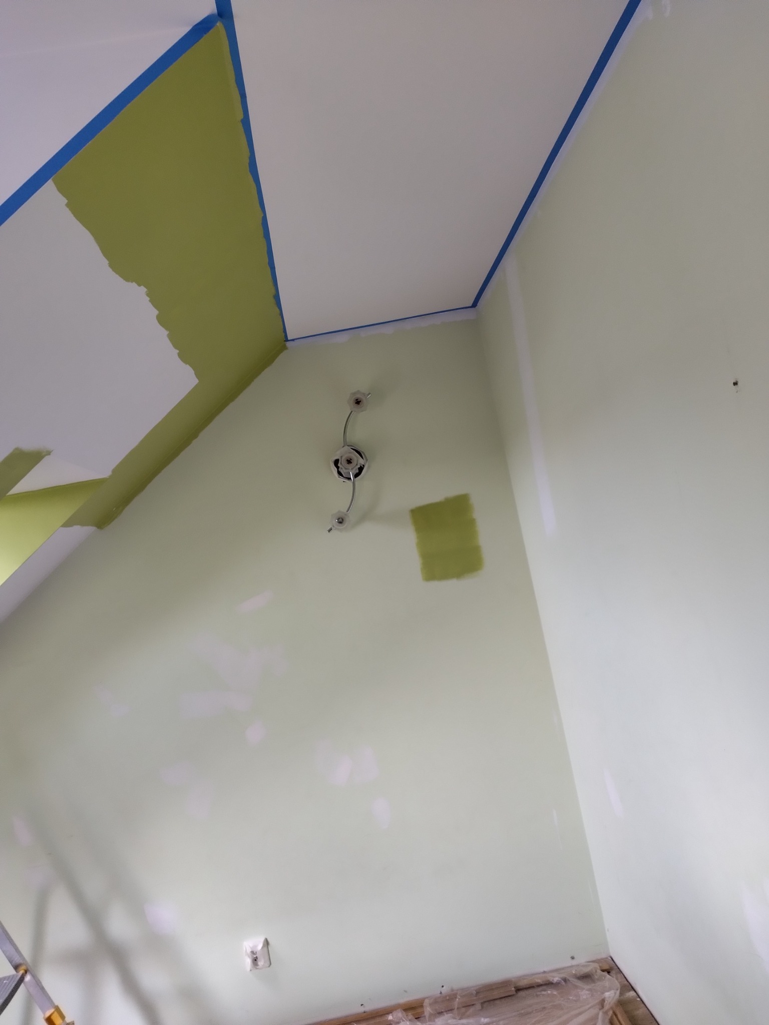 Ściana i fragment sufitu w trakcie malowania na zielono, zabezpieczone taśmą malarską, widoczna instalacja elektryczna oświetleniowa