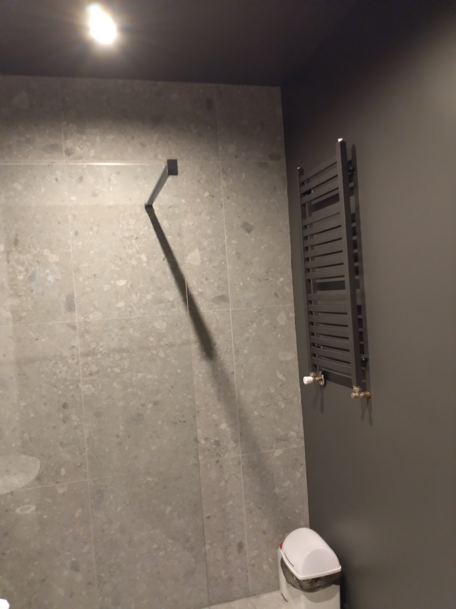 Szary grzejnik drabinkowy zamontowany na ciemnej ścianie, obok ściany wyłożonej płytkami imitującymi kamień, z widocznym koszem na śmieci w dolnym rogu.