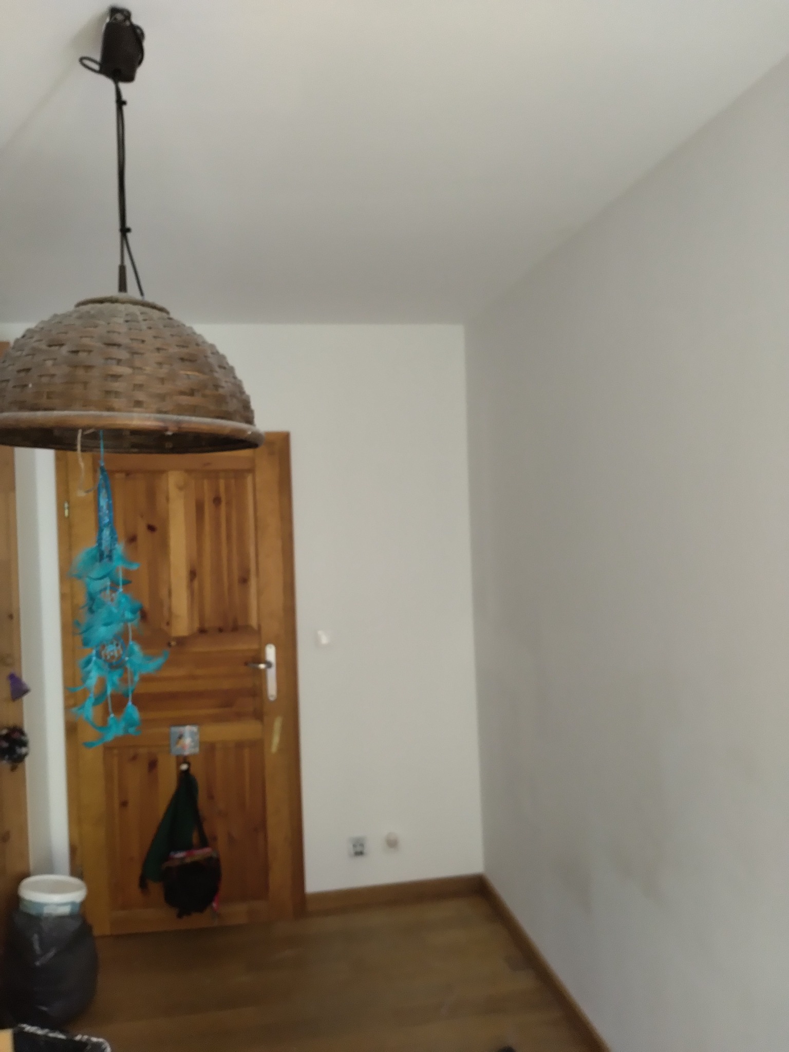 Wnętrze pokoju z drewnianą lampą wiszącą z abażurem w stylu boho i drewnianymi drzwiami w tle, ściany pomalowane na biało.