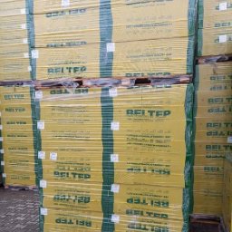 Tani Skład Budowlany - Palety z żółtymi paczkami materiałów izolacyjnych Beltep, owinięte folią stretch, składowane w magazynie.