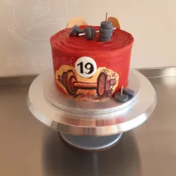 Czerwony tort z kremem, ozdobiony motywem siłowni: sztangą, ciężarami i numerem 19, umieszczony na srebrnej paterze.