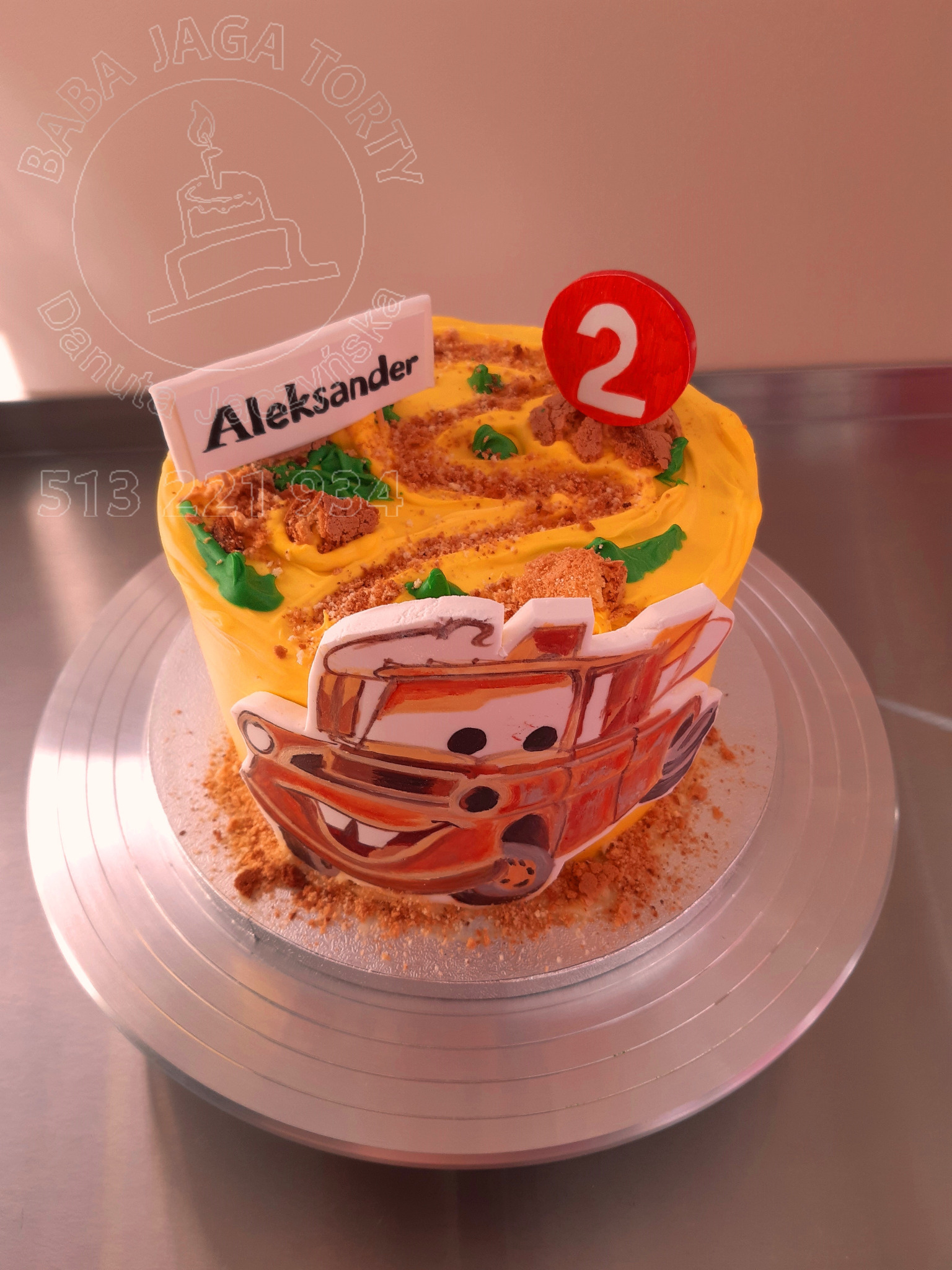 Tort urodzinowy z żółtym kremem, dekoracją z postacią Zygzaka McQueen i napisem 'Aleksander' na białej tabliczce, umieszczony na srebrnej paterze.
