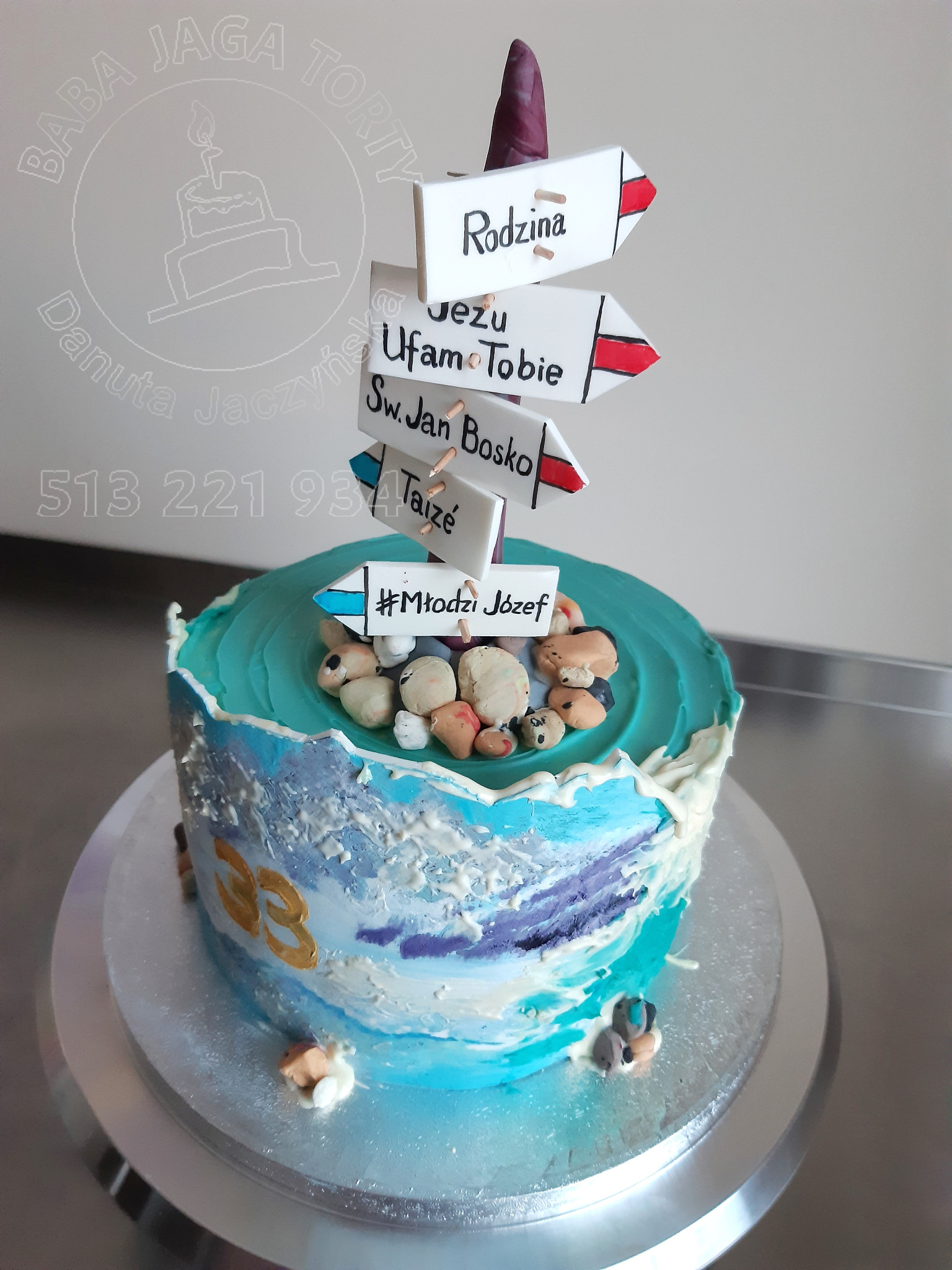 Artystyczny tort z kremem w odcieniach błękitu, fioletu i bieli, udekorowany jadalnymi kamieniami i drogowskazami z napisami: Rodzina, Jezu Ufam Tobie, Św. Jan Bosko, Taize, #Młodzi Józef. Tort...