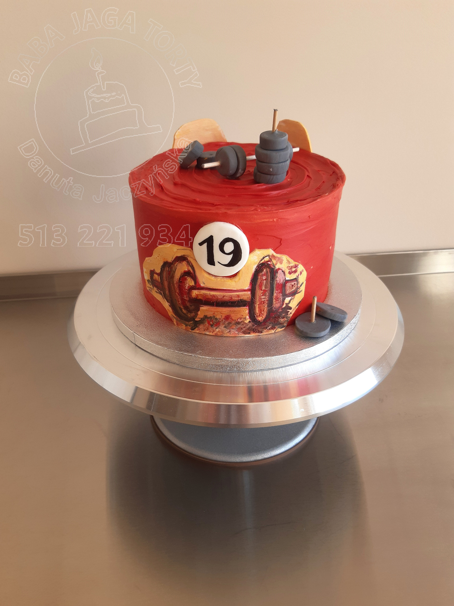 Czerwony tort z kremem, ozdobiony motywem siłowni: sztangą, ciężarami i numerem 19, umieszczony na srebrnej paterze.