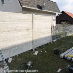 Montaż ogrodzenia betonowego z prefabrykatów: widoczne dwa panele z fakturą cegły, słupki, przygotowane elementy, narzędzia i wiaderka z zaprawą na trawniku, w tle dom i ogrodzenie z siatki.